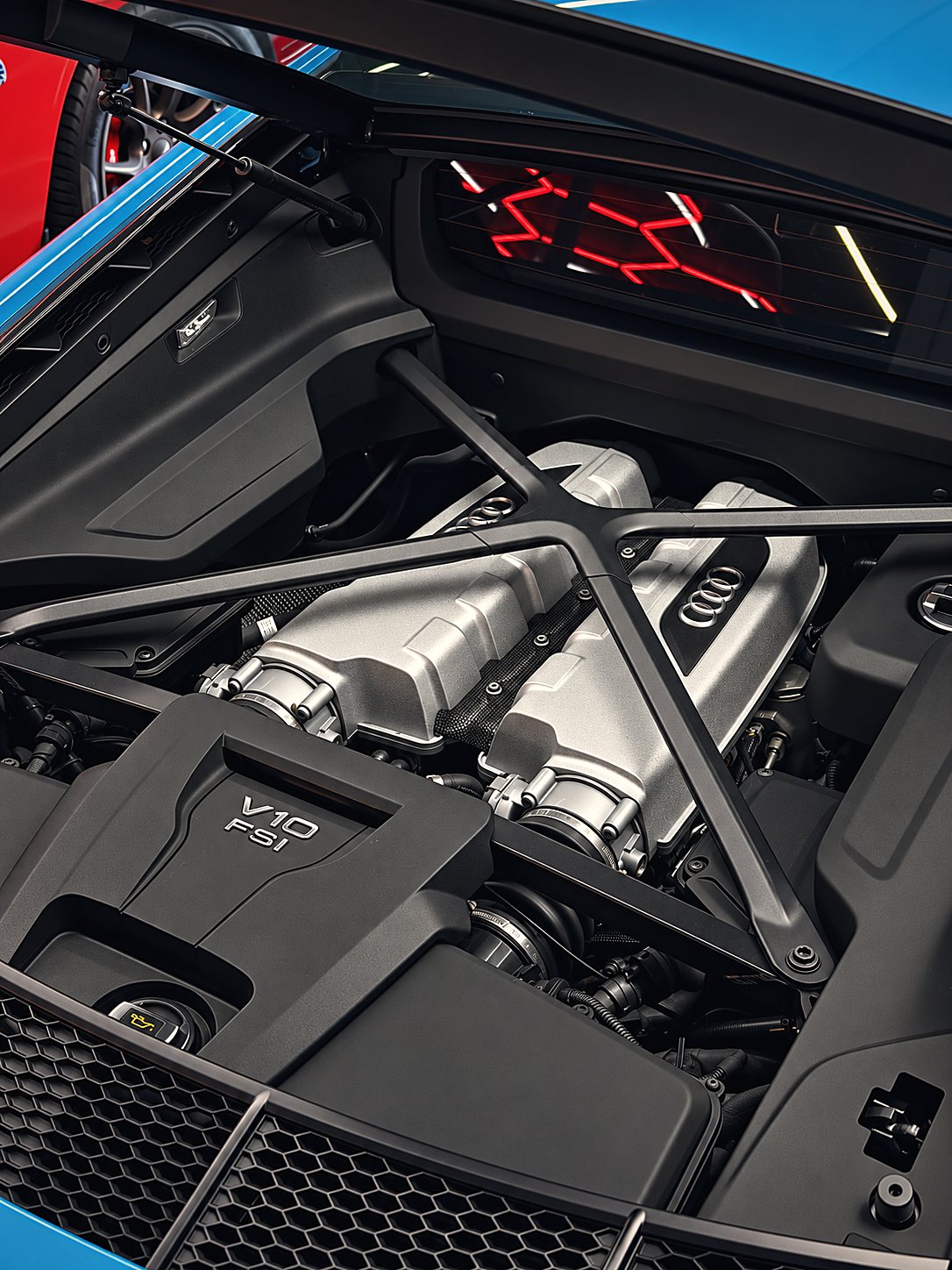 The V10 under the glass of a blue R8 on the club floor, the hex ceiling lights reflected in the window.